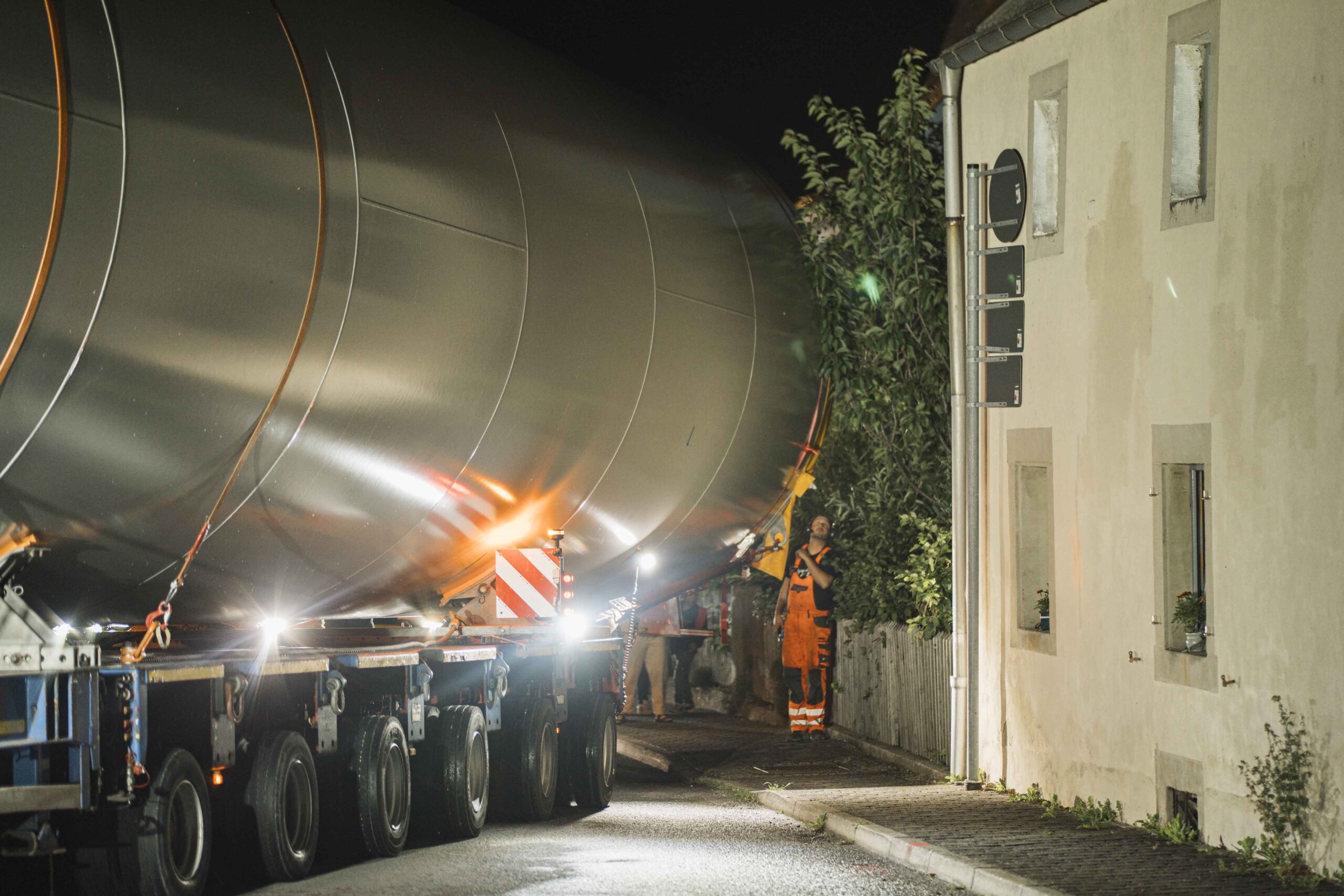 Große Komponenten eines Windrads werden nachts durch eine enge Straße gefahren, ein Arbeiter dirigiert.